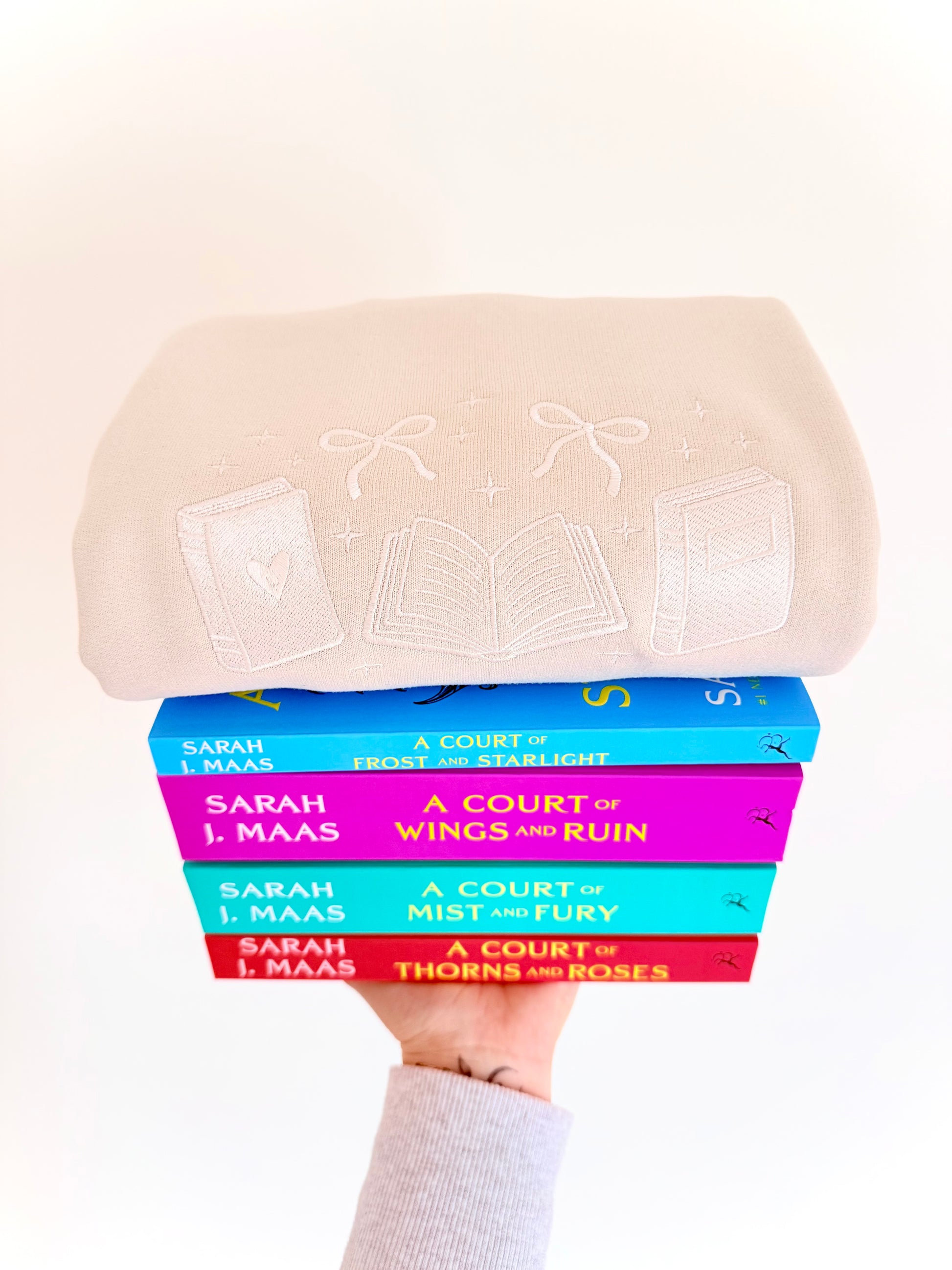 Stack of books by Sarah J. Maas held under a beige hat with lace details.
