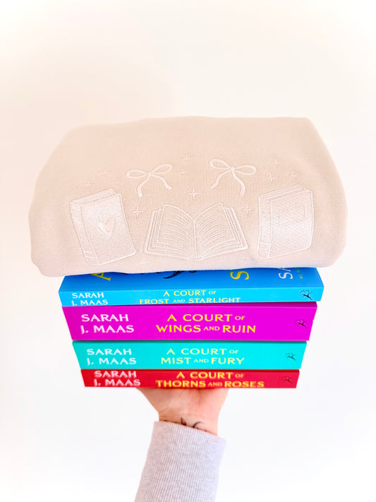 Stack of books by Sarah J. Maas held under a beige hat with lace details.