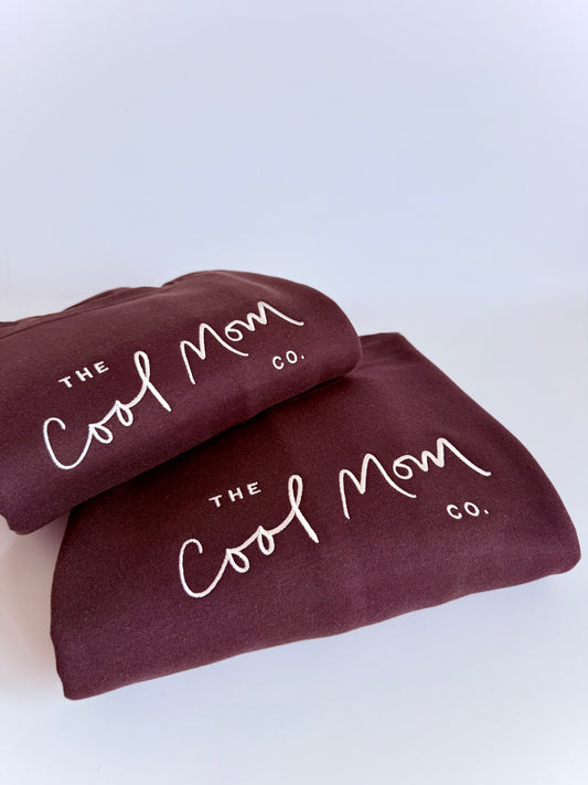 Two brown sweatshirts with 'The Cool Mom Co.' text on a light blue background