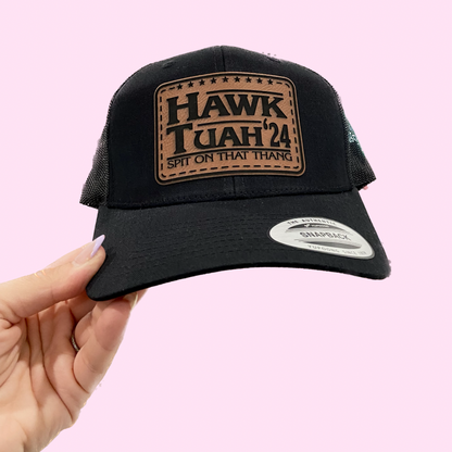 A black snapback hat with a faux leather patch on the front featuring the text 'HAWK TUAH' in a laser engraved design.