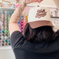 Person wearing a cap with 'Well Sheeple' text, making a heart shape with their hands in front of a wall of colorful bottles.