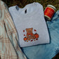 A folded light heather gray sweatshirt with a teddy bear and pumpkin graphic, styled on blue jeans with a plaid blanket and a jar of caramel apple on grass.