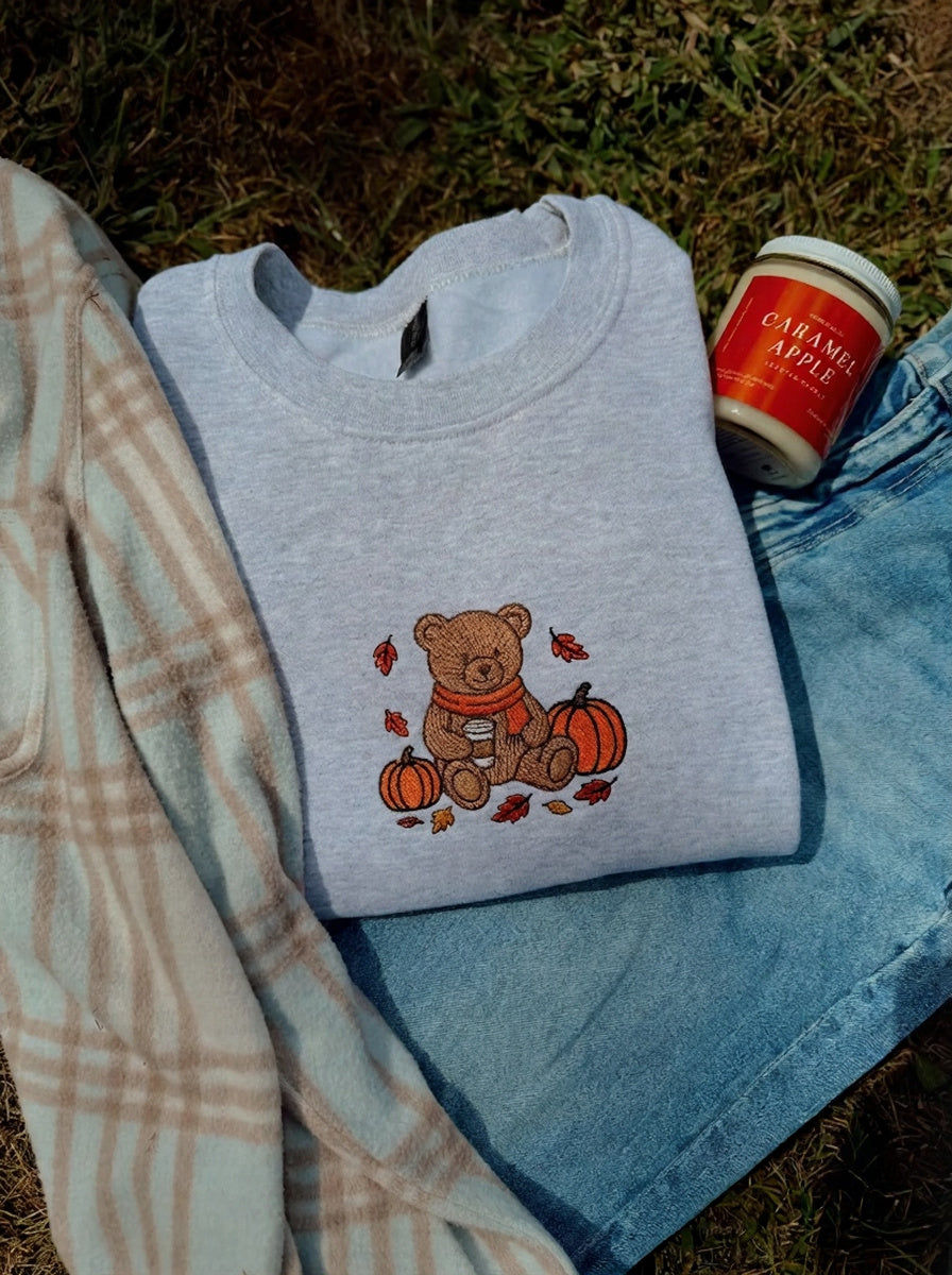 A folded light heather gray sweatshirt with a teddy bear and pumpkin graphic, styled on blue jeans with a plaid blanket and a jar of caramel apple on grass.