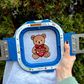 Embroidery hoop with a teddy bear design held by a person outdoors.
