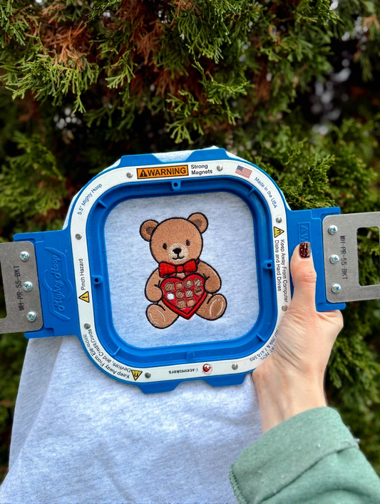Embroidery hoop with a teddy bear design held by a person outdoors.